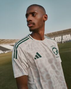 Portrait of a man in a white Saudi football jersey standing on a grassy field with a stadium in the background, looking off to the side.