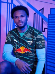 Young man seated in a sports jersey with a Red Bull logo, posing casually in a blue-lit backdrop with geometric patterns.