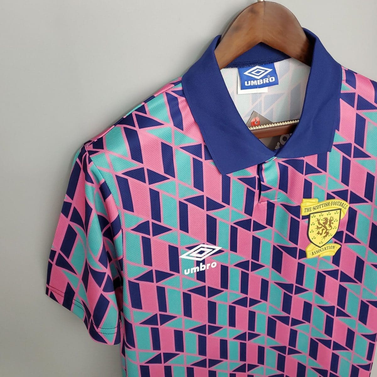 Close-up of a colorful geometric-patterned soccer jersey on a wooden hanger, featuring a blue collar, the Umbro logo, and the Scottish Football Association badge.