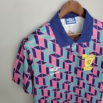 Close-up of a colorful geometric-patterned soccer jersey on a wooden hanger, featuring a blue collar, the Umbro logo, and the Scottish Football Association badge.