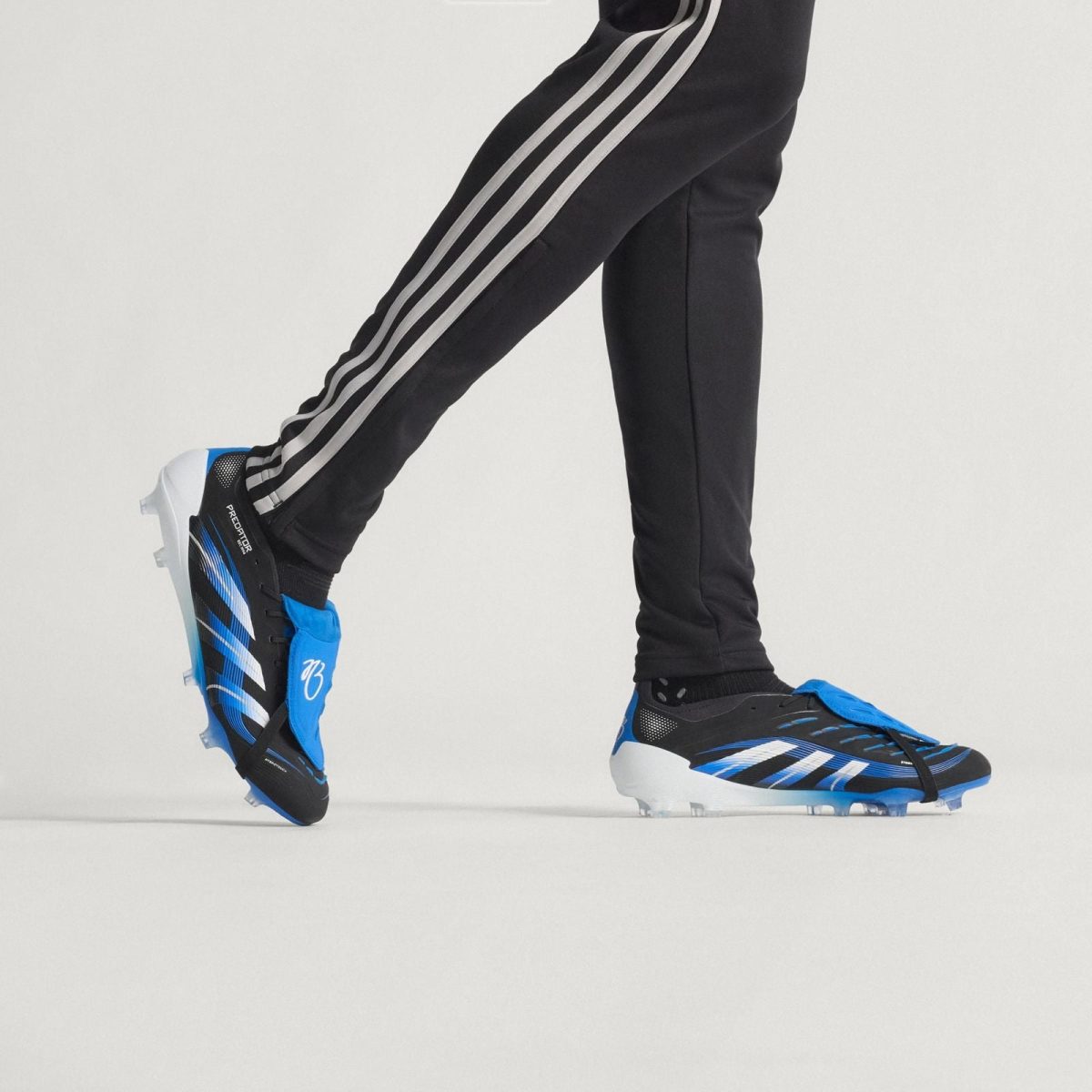 Person wearing black athletic pants with white side stripes, stepping in blue and black cleats on a light background