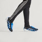 Person wearing black athletic pants with white side stripes, stepping in blue and black cleats on a light background