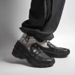ea78ef7f-6456-4c57-b2f2-2c3c6ad78dd1 | UAE SPORT STORE Close-up of black chunky sneakers on a person’s feet, light socks visible, pant legs cuffed against a gray backdrop.