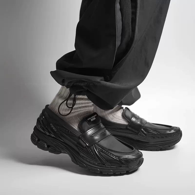 ea78ef7f-6456-4c57-b2f2-2c3c6ad78dd1 | UAE SPORT STORE Close-up of black chunky sneakers on a person’s feet, light socks visible, pant legs cuffed against a gray backdrop.