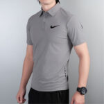 Man wearing a gray short-sleeve polo with a black Nike swoosh on the chest, standing against a light wall.
