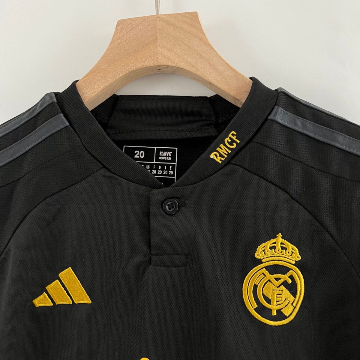 Black jacket on a wooden hanger with a yellow Real Madrid crest embroidered on the chest and yellow Adidas stripes visible on the left shoulder area.