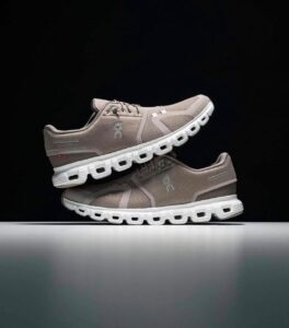Pair of beige mesh running shoes with white soles stacked one atop the other against a dark background, showcasing textured upper and cutout sole design.