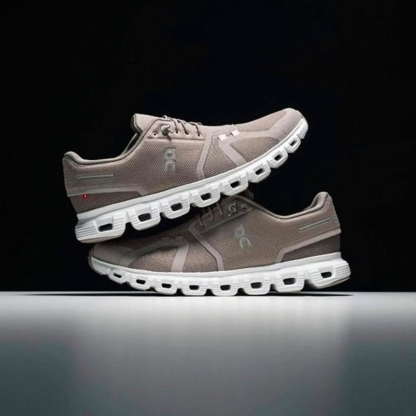 Pair of beige mesh running shoes with white soles stacked one atop the other against a dark background, showcasing textured upper and cutout sole design.