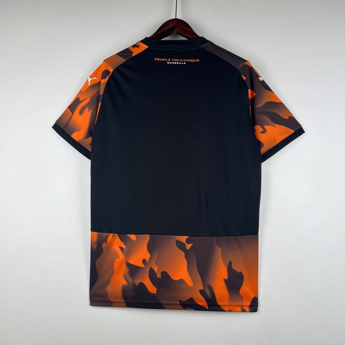 Back view of a black T-shirt with orange camouflage sleeves hanging on a wooden hanger; text 'PEUPLE VOLCANIQUE MARSEILLE' visible on the upper back.