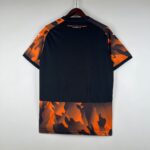eab613c2_38366d8d-655d-4f19-8b84-5280fc1f0873 | UAE SPORT STORE Back view of a black T-shirt with orange camouflage sleeves hanging on a wooden hanger; text 'PEUPLE VOLCANIQUE MARSEILLE' visible on the upper back.
