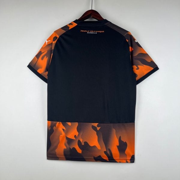 Back view of a black T-shirt with orange camouflage sleeves hanging on a wooden hanger; text 'PEUPLE VOLCANIQUE MARSEILLE' visible on the upper back.