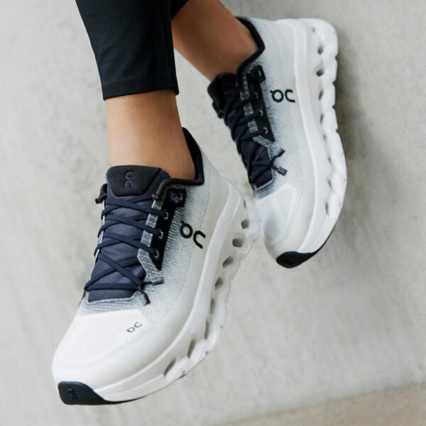 White and gray On running shoes with black laces worn by a person in black leggings, standing on a concrete surface.