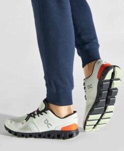 Person wearing white On running shoes with orange heel grips and black sole, standing in navy joggers.