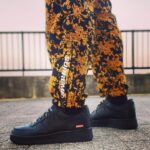 Black Nike Air Force 1 sneakers with a red Supreme tag, paired with yellow floral-pattern pants on a paved surface.