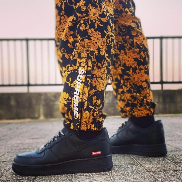 Black Nike Air Force 1 sneakers with a red Supreme tag, paired with yellow floral-pattern pants on a paved surface.