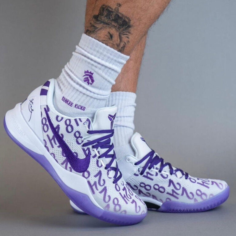 White and purple basketball sneakers with a number-pattern upper, purple laces and sole, worn with white crew socks.