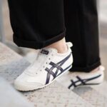 Close-up of white sneakers with blue stripes stepping up onto a metal grate, worn with black rolled-up pants.