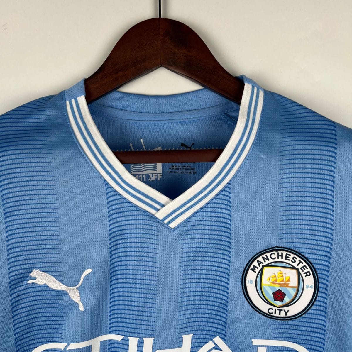 Blue Manchester City soccer jersey on a wooden hanger, close-up of the collar and crest visible on the right side of the image.
