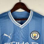 Blue Manchester City soccer jersey on a wooden hanger, close-up of the collar and crest visible on the right side of the image.