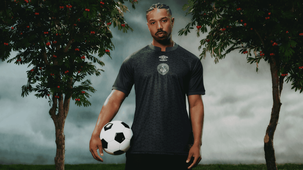Male soccer player in a dark jersey holds a ball at his side between two fruit trees with red berries in a moody outdoor setting.