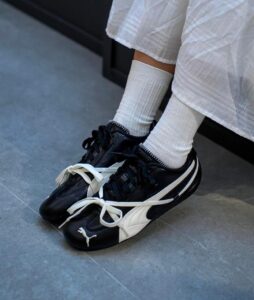 Person wearing black and white Puma sneakers with white socks and a white dress, standing on a gray floor.
