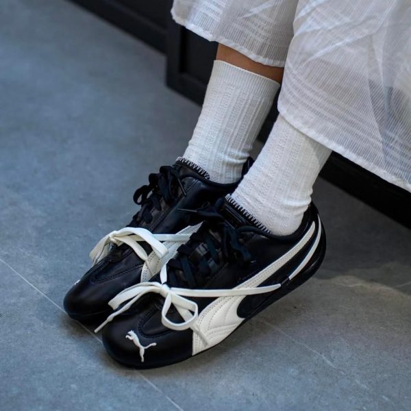 Person wearing black and white Puma sneakers with white socks and a white dress, standing on a gray floor.