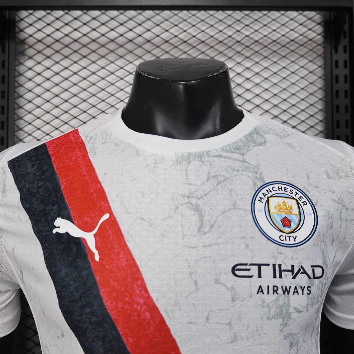 Close-up of a Manchester City football jersey on a black mannequin, featuring the crest and a red-blue diagonal stripe against a mesh backdrop.