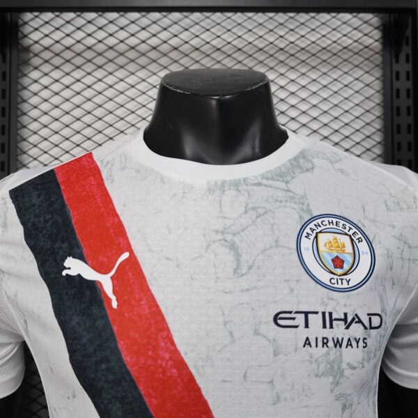 Close-up of a Manchester City football jersey on a black mannequin, featuring the crest and a red-blue diagonal stripe against a mesh backdrop.