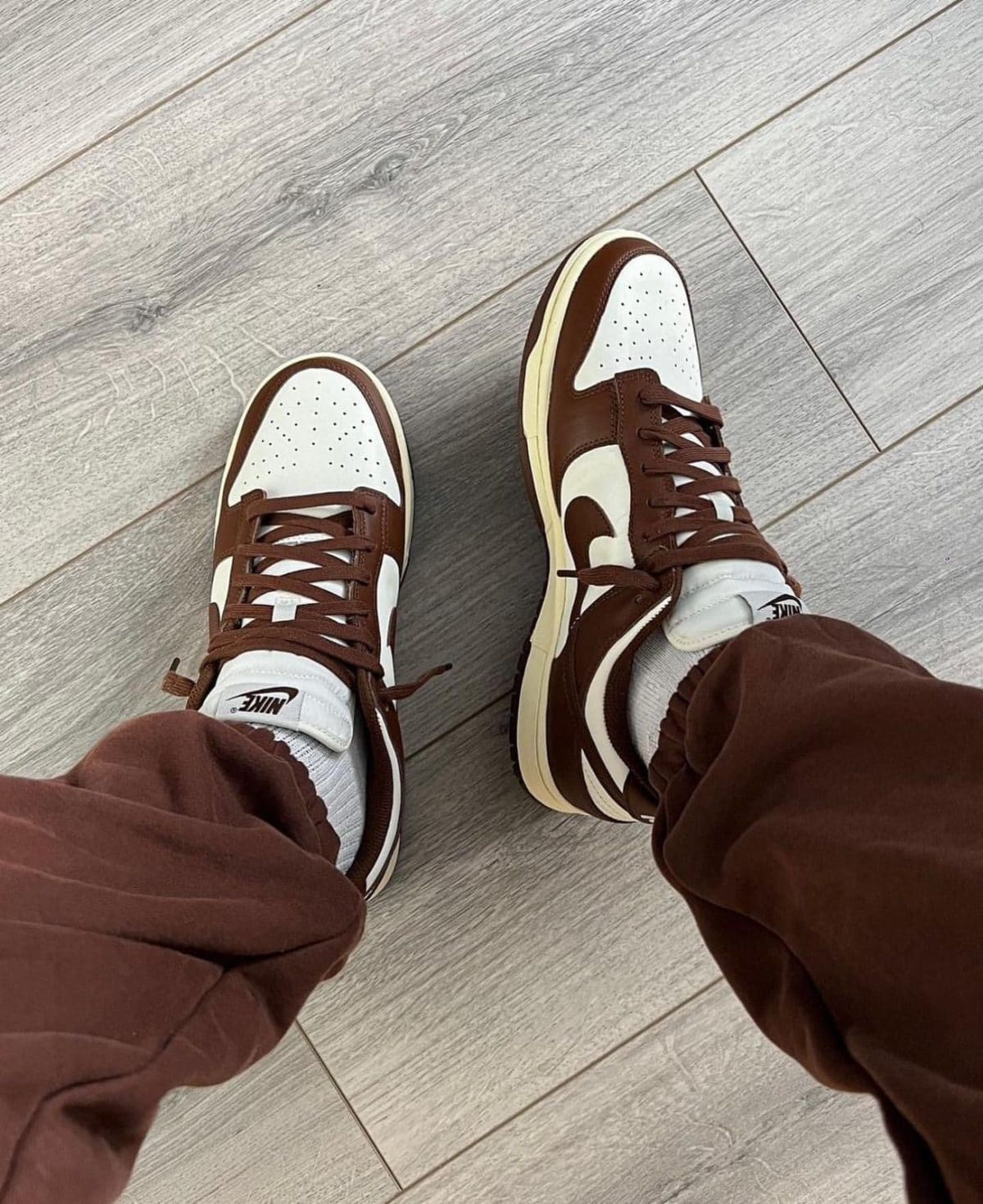 Top-down view of brown and white Nike sneakers on a light wood floor, with brown pants visible at the ankles.
