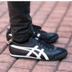 Close-up of black sneakers with white stripes worn on a brick-paved walkway.