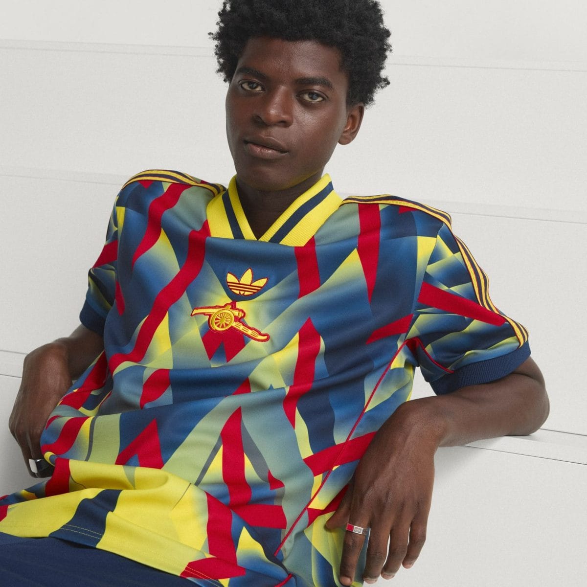 Young man with dark skin wearing a vibrant geometric Adidas jersey, sitting against a light background, facing the camera.