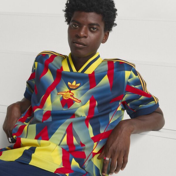 Young man with dark skin wearing a vibrant geometric Adidas jersey, sitting against a light background, facing the camera.