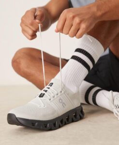 Person ties white athletic shoes, focusing on lacing the sneaker with striped socks visible in the background as they tie the laces
