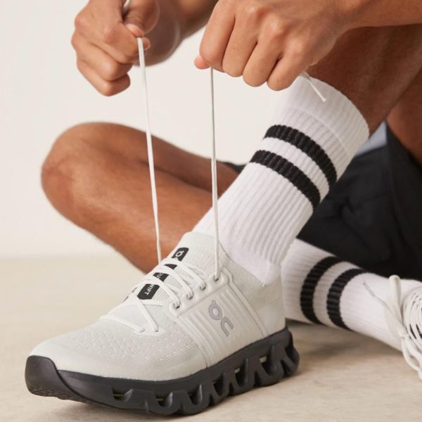 Person ties white athletic shoes, focusing on lacing the sneaker with striped socks visible in the background as they tie the laces