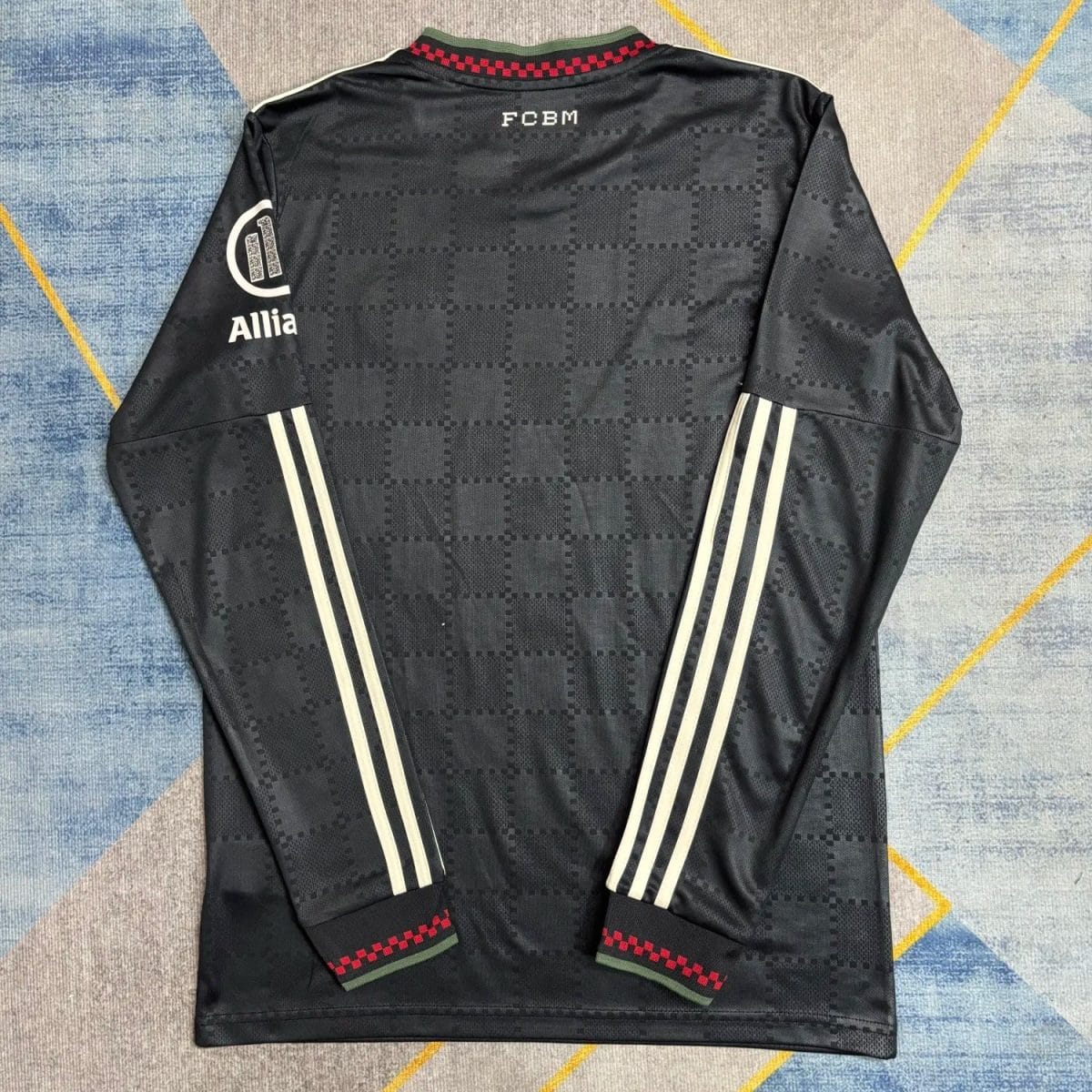 Back view of a black training jersey with a subtle checkerboard pattern, white triple stripes along each sleeve, and red-green cuffs at the hem. FCBM text appears on the upper back with an Allia logo on the left sleeve.