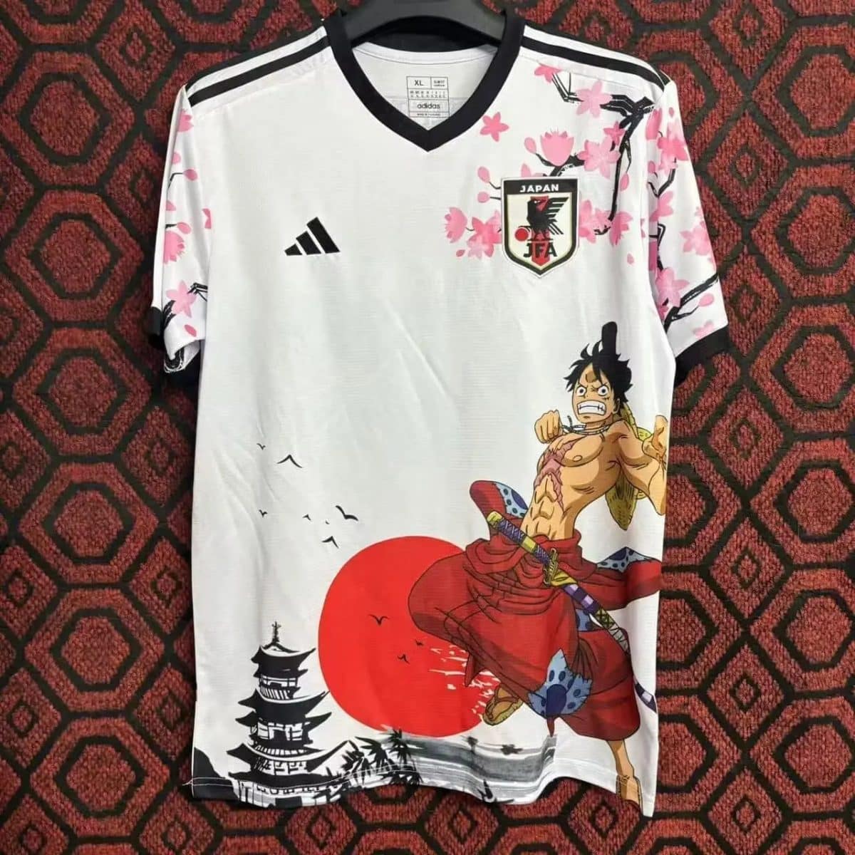 White Japan soccer jersey with black V-neck, Adidas logo, pink sakura blossoms, and a large Luffy from One Piece illustration on the front.
