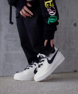 Person in black jeans and a colorful-sleeve hoodie wears white Nike sneakers with a black swoosh against a concrete wall.