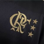 Gold embroidered monogram logo with curved interlaced letters and three gold stars on dark textured fabric, likely on a jacket.