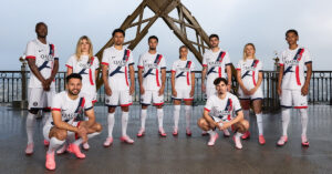 Eleven soccer players in PSG white kits with Qatar Airways logos stand in a row on a rooftop with a metal structure behind them, posing for a team photo.