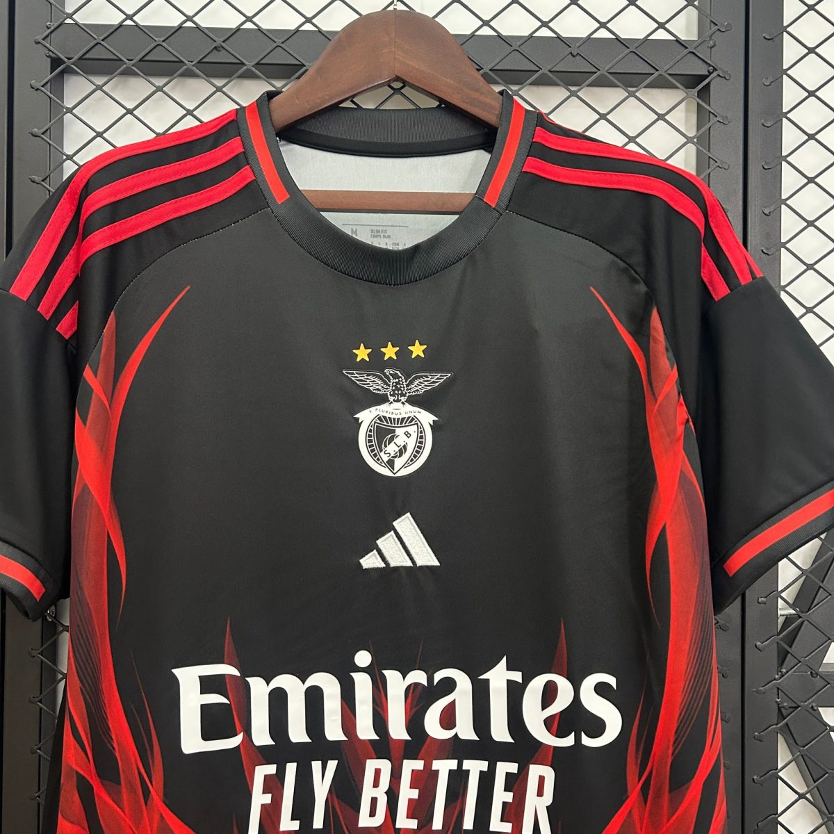 Black football jersey on hanger with red stripes, crest, three gold stars, and 'Emirates Fly Better' slogan visible