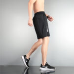 Side view of a shirtless man walking in black athletic shorts and black Nike sneakers, against a gray wall.