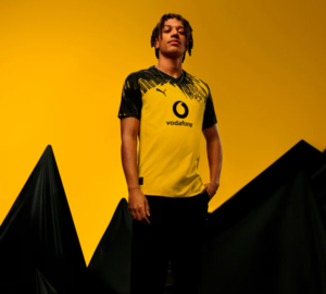 Young man in Borussia Dortmund jersey (yellow and black) stands confidently against a bright yellow backdrop, Vodafone sponsor visible on chest.