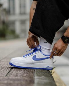 Person tying the blue-lace, white Nike sneaker on a curb, wearing white socks and black pants.