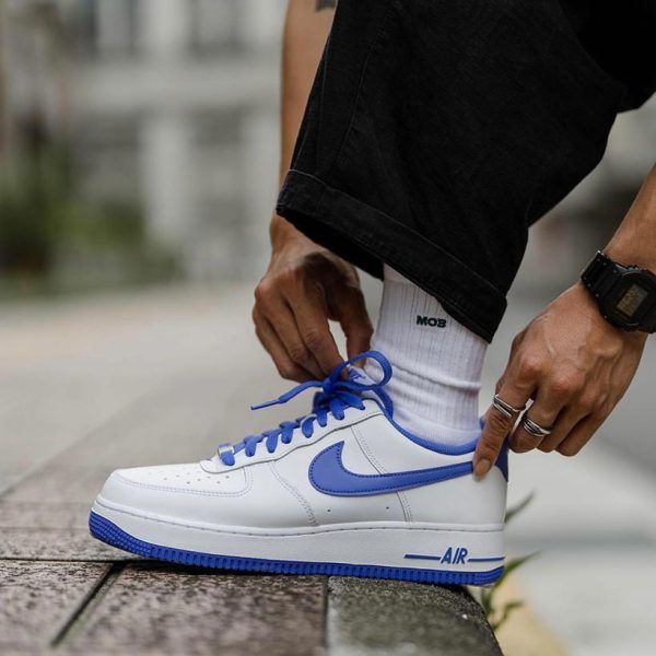 Person tying the blue-lace, white Nike sneaker on a curb, wearing white socks and black pants.