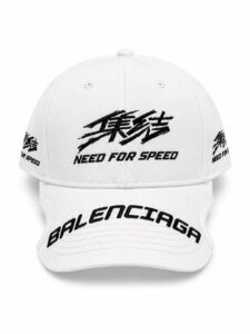 White baseball cap with black embroidery: front features stylized characters and 'NEED FOR SPEED'; curved 'BALENCIAGA' text on the brim; additional small text and logos on the sides.