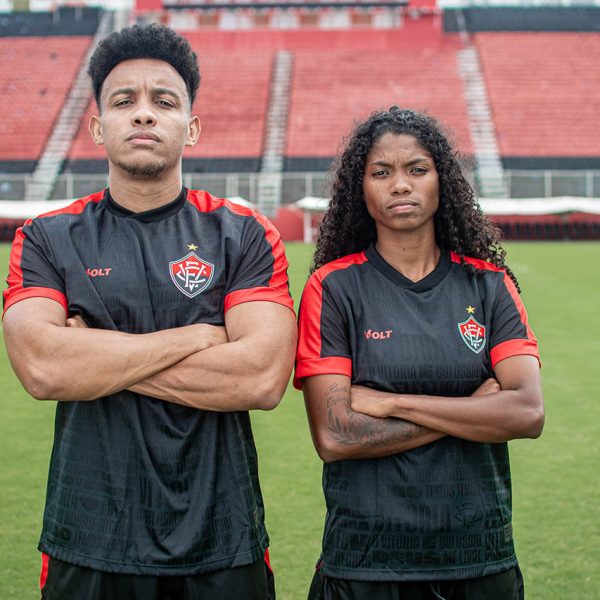 Two soccer players with arms crossed on a green pitch, wearing black and red jerseys, in a stadium.