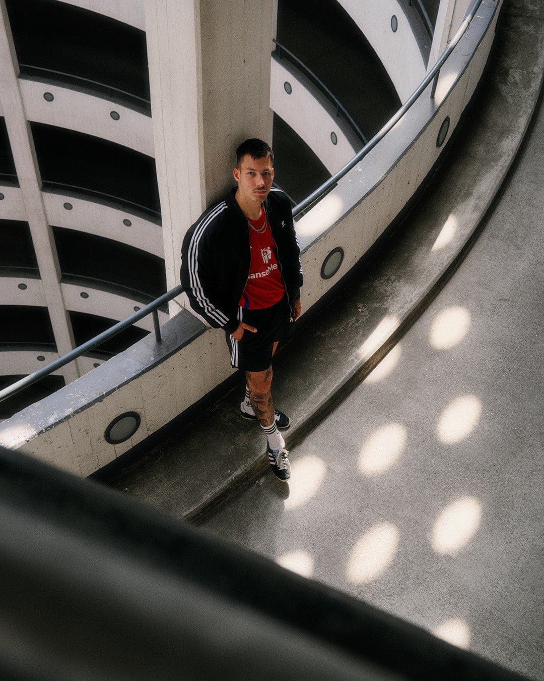 er1SAaf86lCenQc | UAE SPORT STORE Man in a black Adidas tracksuit and red shirt stands on a curved concrete ramp inside a multi-level parking garage, looking at the camera.
