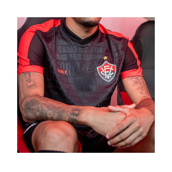 Close-up of a person in a black-red soccer jersey with a shield crest, hands clasped, showing arm tattoos.