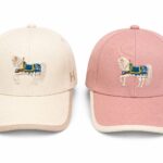 Cream and pink baseball caps with an embroidered white horse and blue saddle on the front panel, side-by-side.