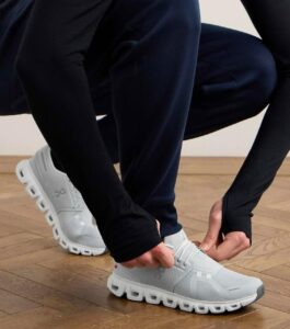 f0f5ad7c-9e83-47f6-99c7-e0fa162ddf5e | UAE SPORT STORE Person in dark athletic wear tying the laces of a light gray sneaker with a chunky sole on a wooden floor